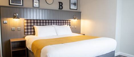 A neatly arranged bedroom with a large bed, a headboard with wall-mounted lights, a small nightstand, and a plaid-patterned rug.