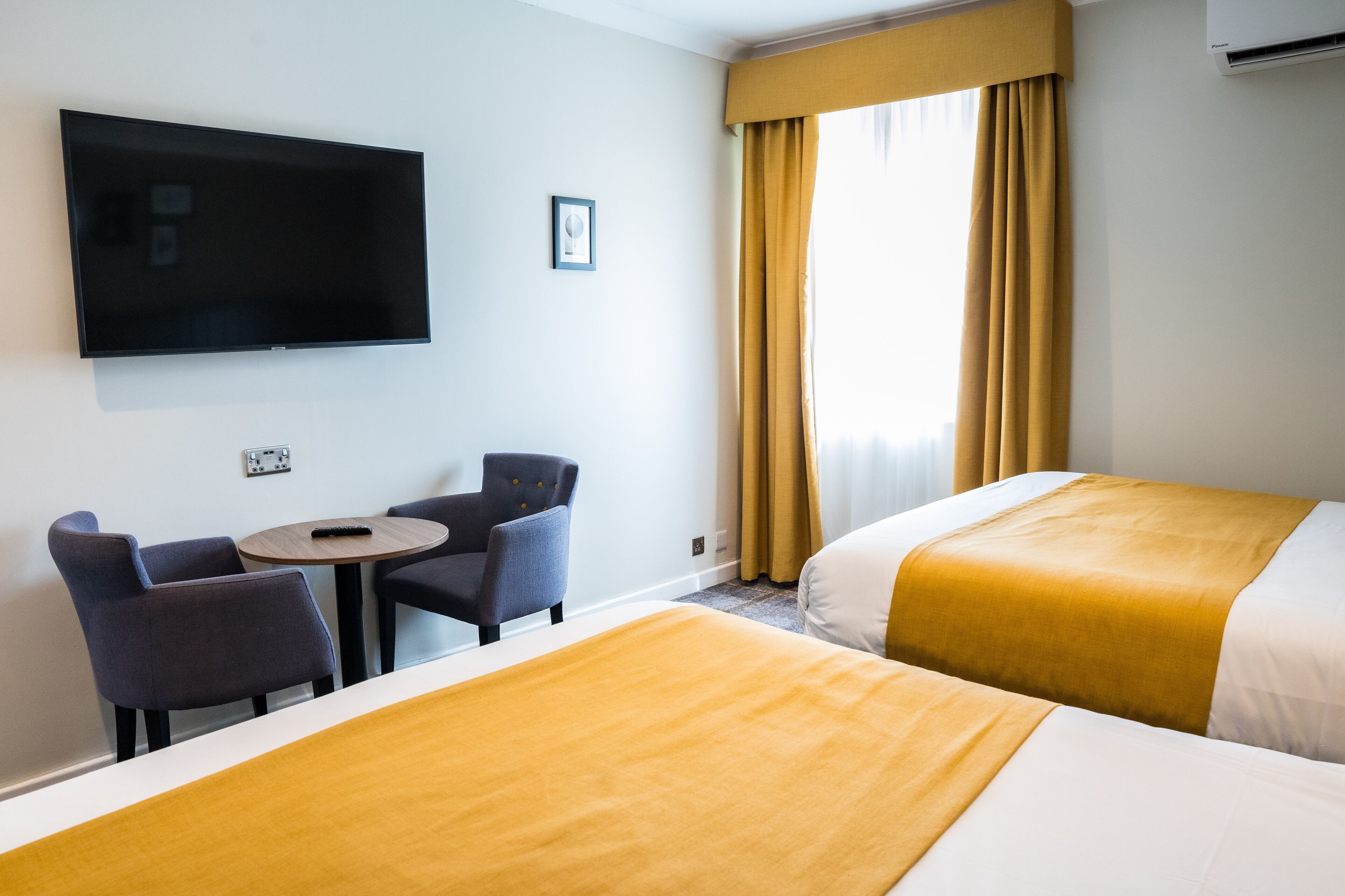 Habitación de hotel con dos camas, televisión, una mesita pequeña con dos sillas y una ventana con cortinas.