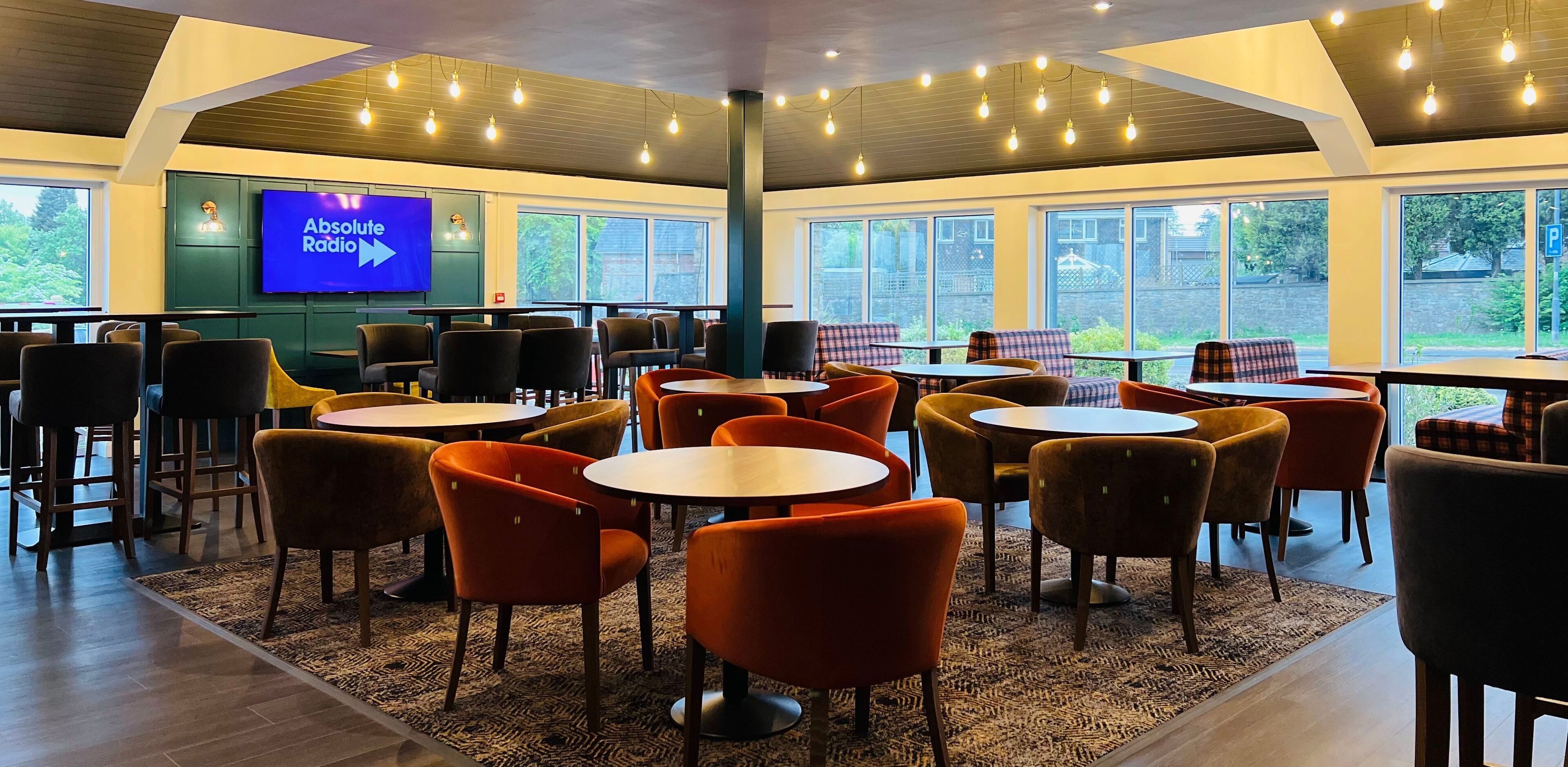 A modern cafe with round tables and orange chairs, a large TV screen displaying 'Absolute Radio', and large windows offering a view of the outdoors.