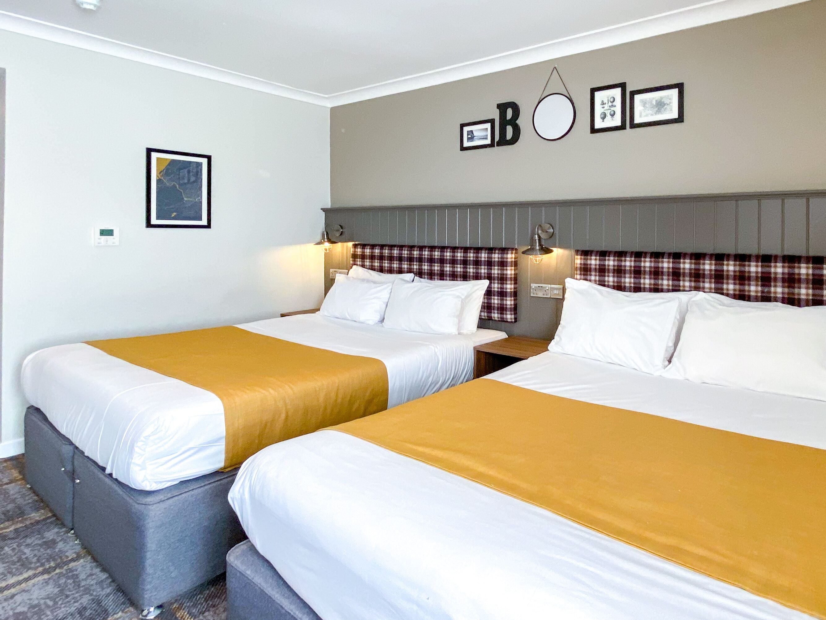 Habitación de hotel con dos camas, cada una con ropa de cama blanca y una manta color mostaza. Hay luces empotradas en la pared y un cuadro enmarcado sobre las camas.