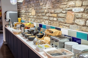 Montaje de bufé con diversos alimentos, una máquina de café y una pared de piedra al fondo.