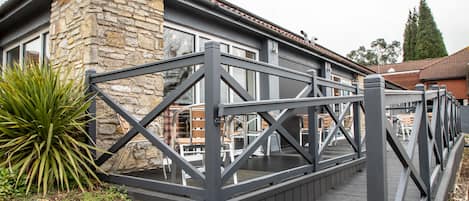 A wooden deck with a metal railing, leading to a stone building with large windows.