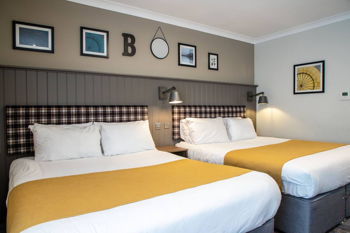 Duas camas com roupa de cama branca e mostarda, uma luminária na parede e quadros emoldurados.