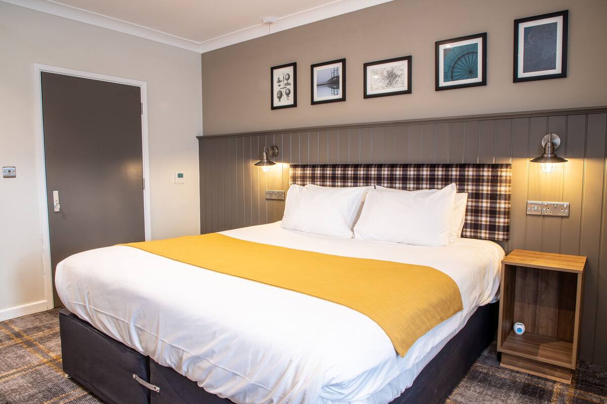 Uma cama bem arrumada com um cobertor amarelo, cabeceira xadrez e mesa de cabeceira com abajur.