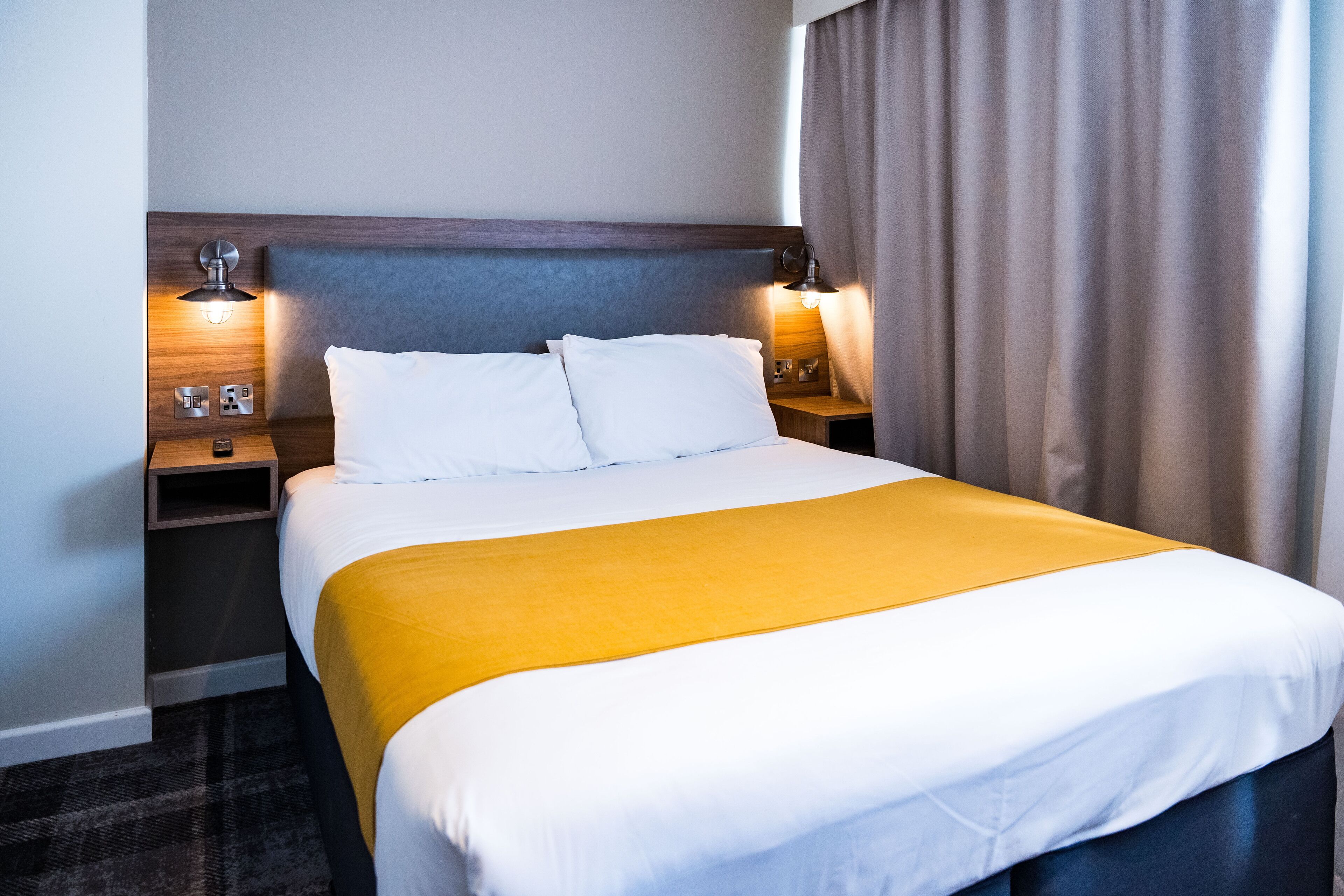 A hotel room with a large bed, two bedside tables with lamps, a wooden headboard, and a window with curtains.