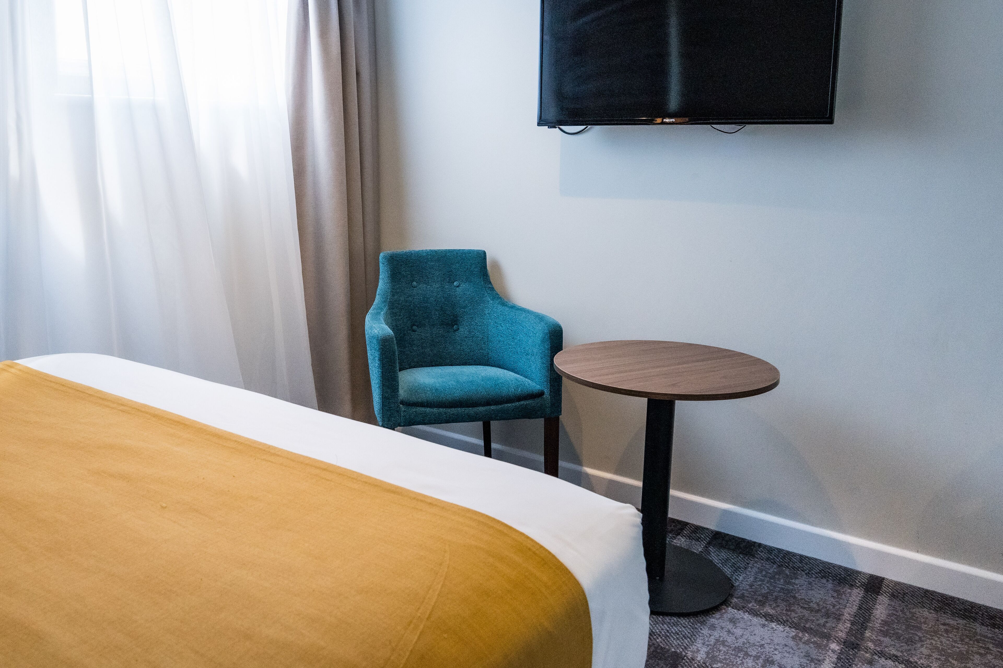 A hotel room with a bed, a blue armchair, a small round table, and a TV mounted on the wall.