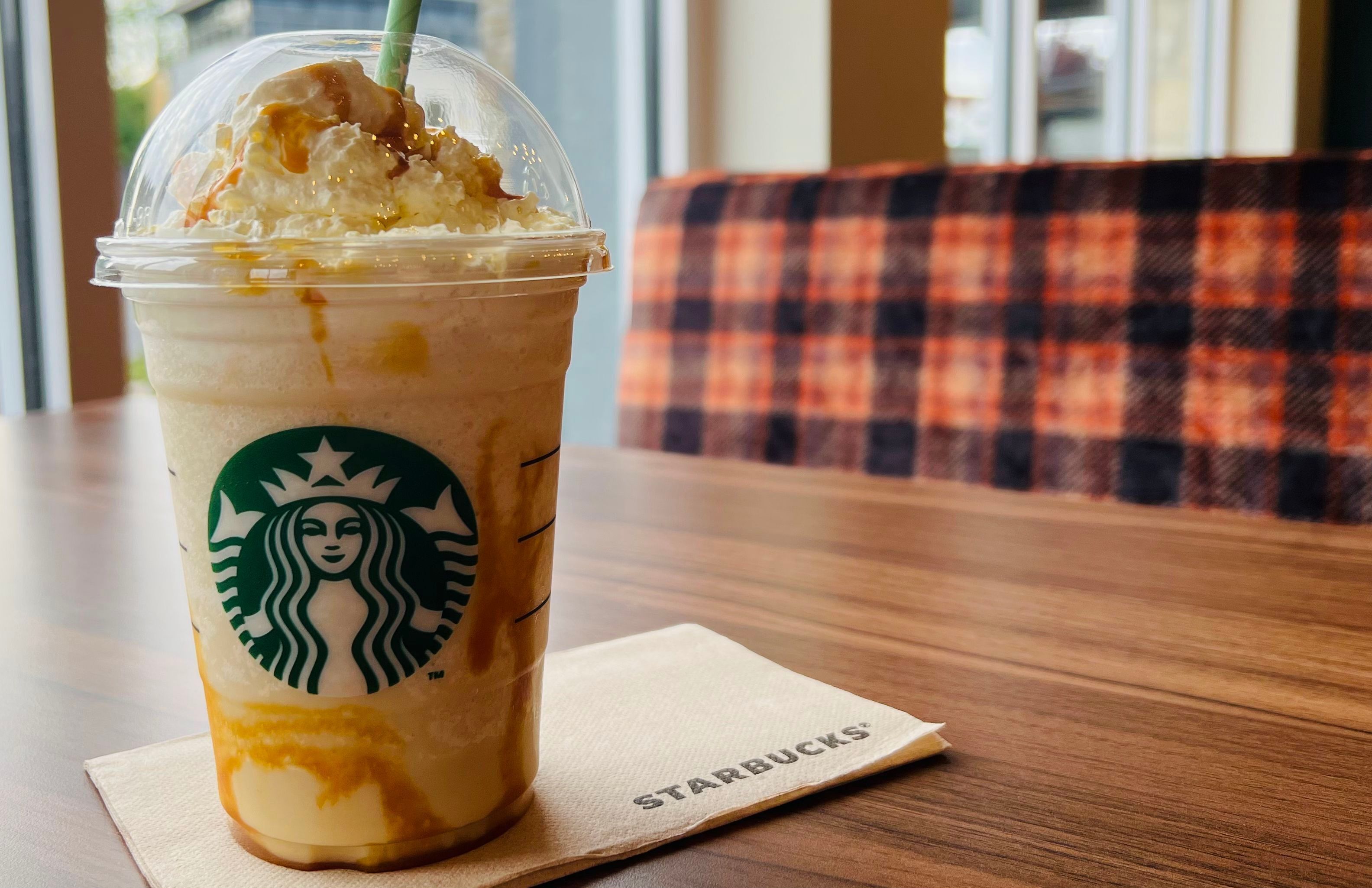 Ένας παγωμένος καφές Starbucks με σαντιγί και σιρόπι καραμέλας, σερβιρισμένος σε ένα διαφανές πλαστικό ποτήρι με καπάκι, τοποθετημένο πάνω σε ένα ξύλινο τραπέζι, με ένα καρό μαξιλάρι στο φόντο.