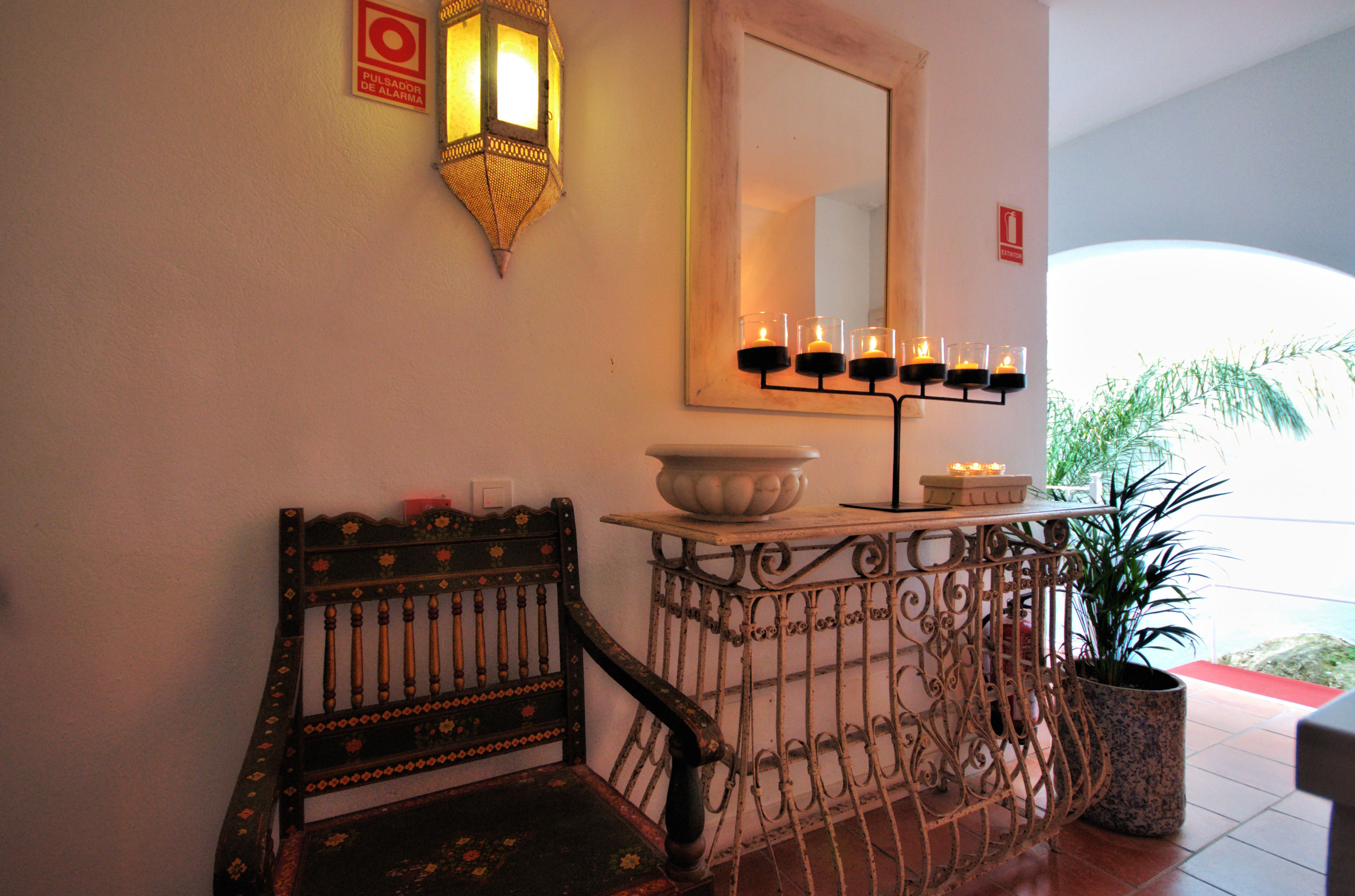 Una habitación con una silla de madera, una mesa con un cuenco y velas, un espejo adosado a la pared y una planta en maceta.