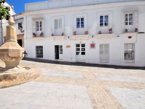 A white building with multiple windows and balconies, a stone fountain in the foreground, and a cobblestone street. - Benali Guesthouse (Tarifa)