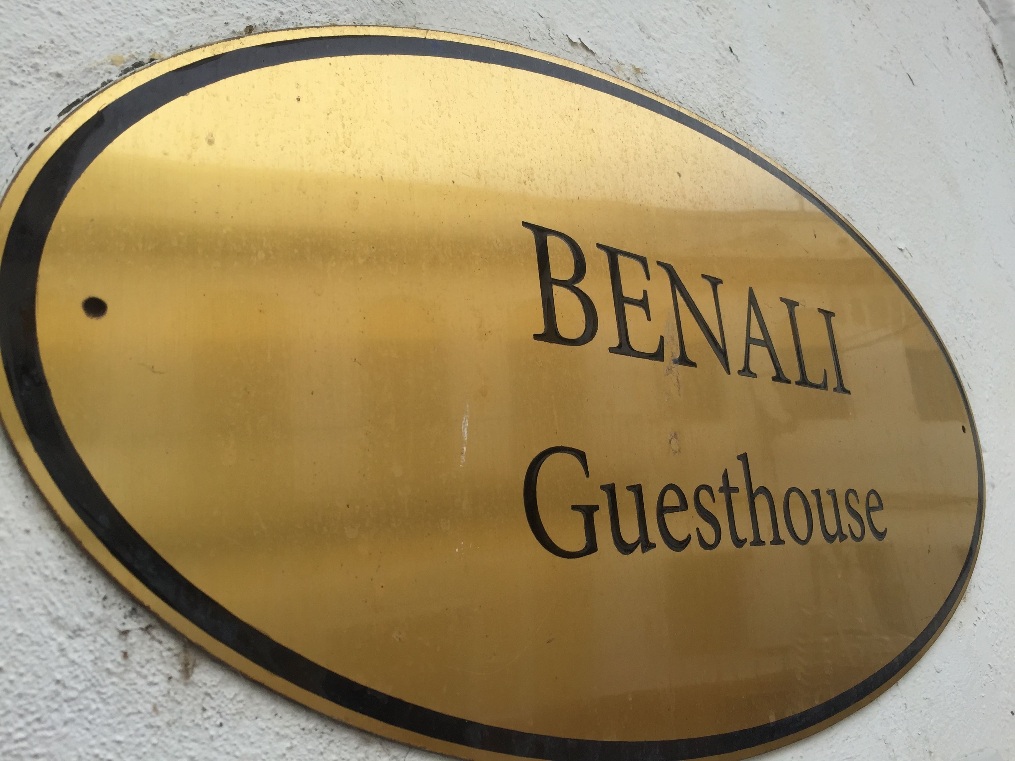 A gold-colored oval sign with the text "BENALI Guesthouse" on a white wall.