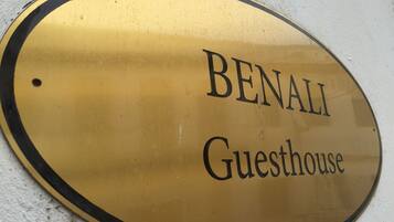 Een ovaal bord in goudkleur met de tekst 'BENALI Guesthouse' op een witte muur.