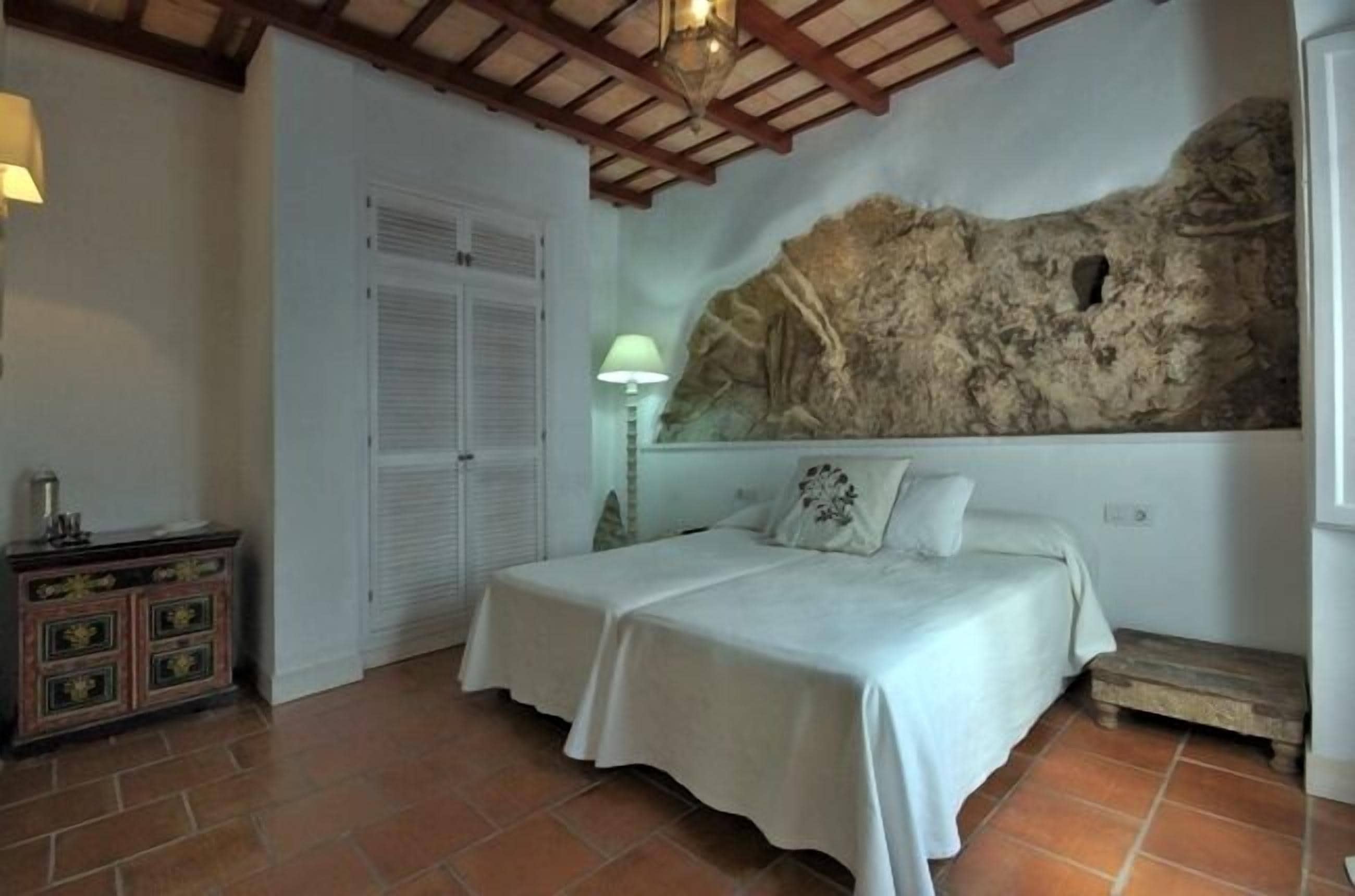 A bedroom with two beds, a stone wall, a wooden ceiling, and a tiled floor.