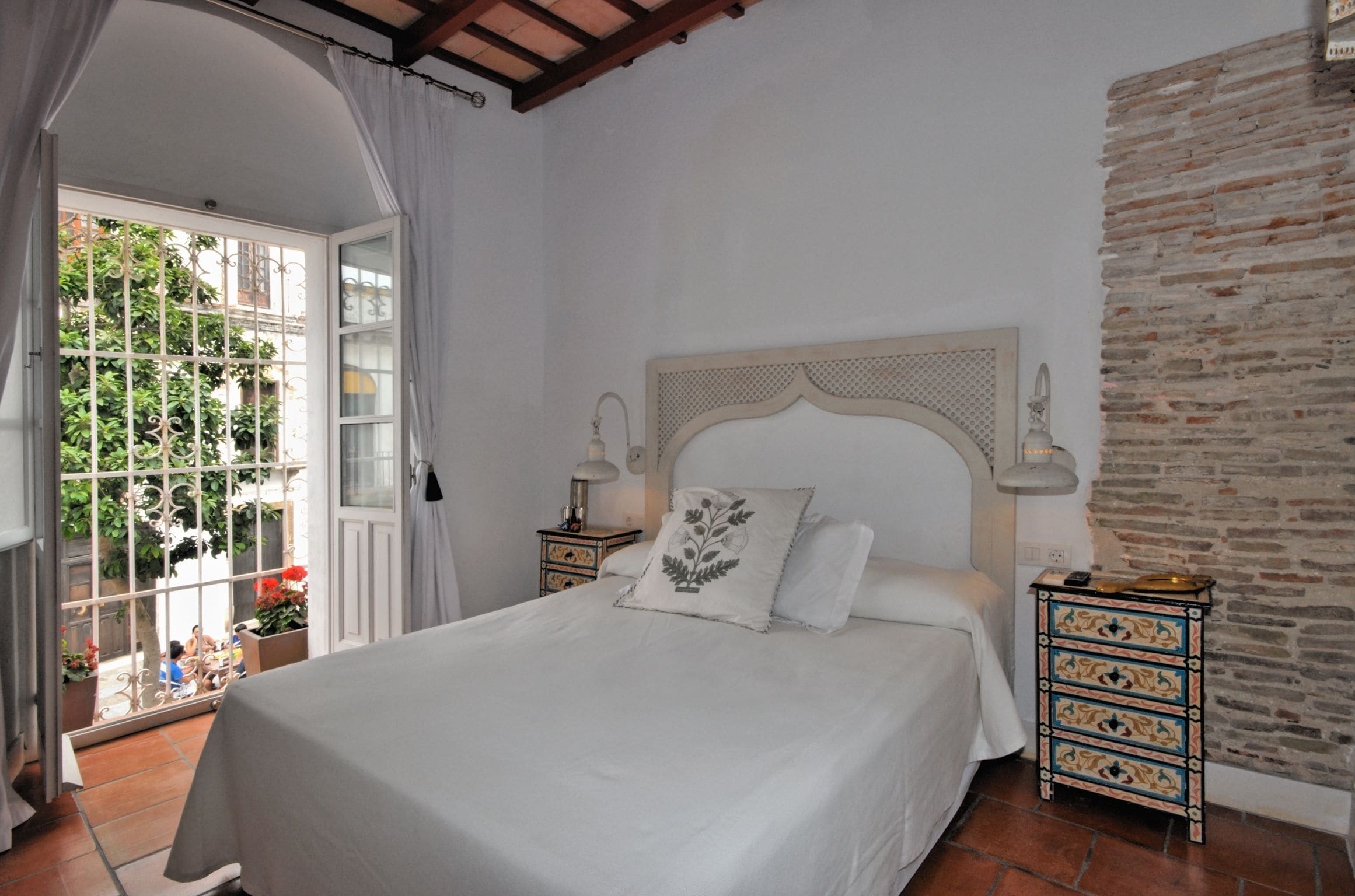 Een slaapkamer met een groot bed, een houten hoofdeinde, een nachtkastje met een lamp en uitzicht op een tuin door een raam.