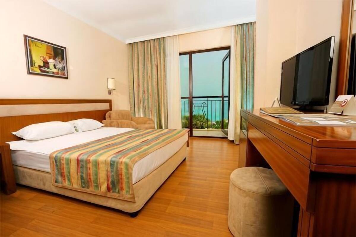A hotel room with a bed, a desk with a television, a window with curtains, and a balcony with a view.