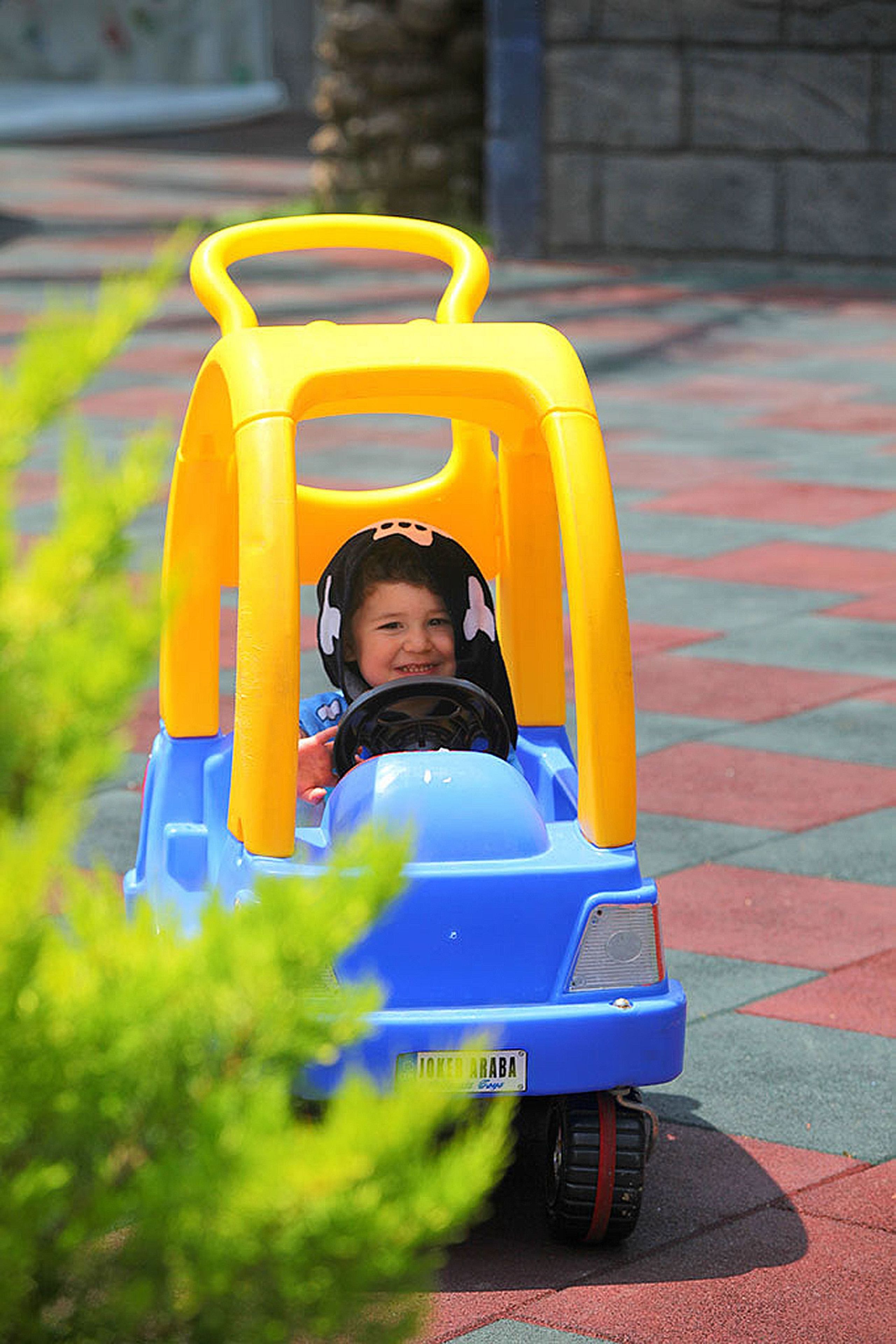 A child in a small, yellow and blue toy car with a steering wheel, driving on a paved surface.
