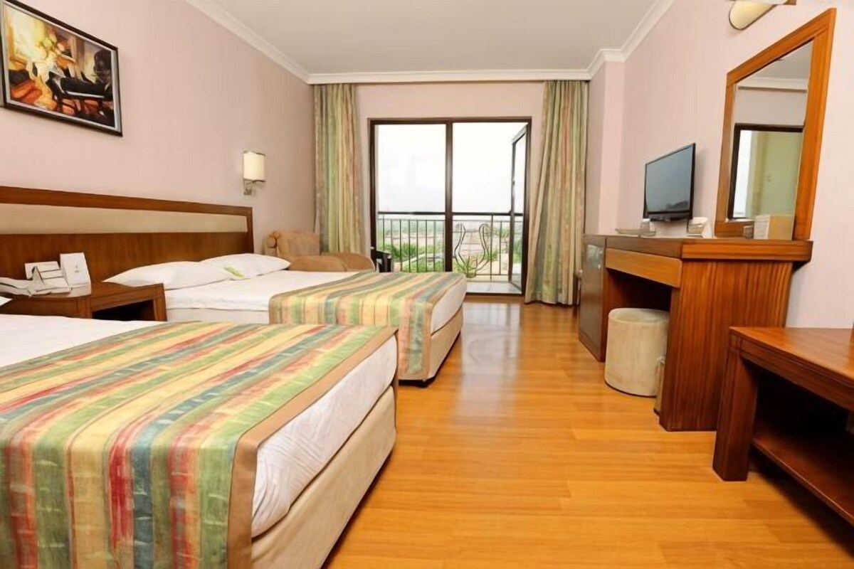 A hotel room with two beds, a desk, a television, and a balcony with curtains.