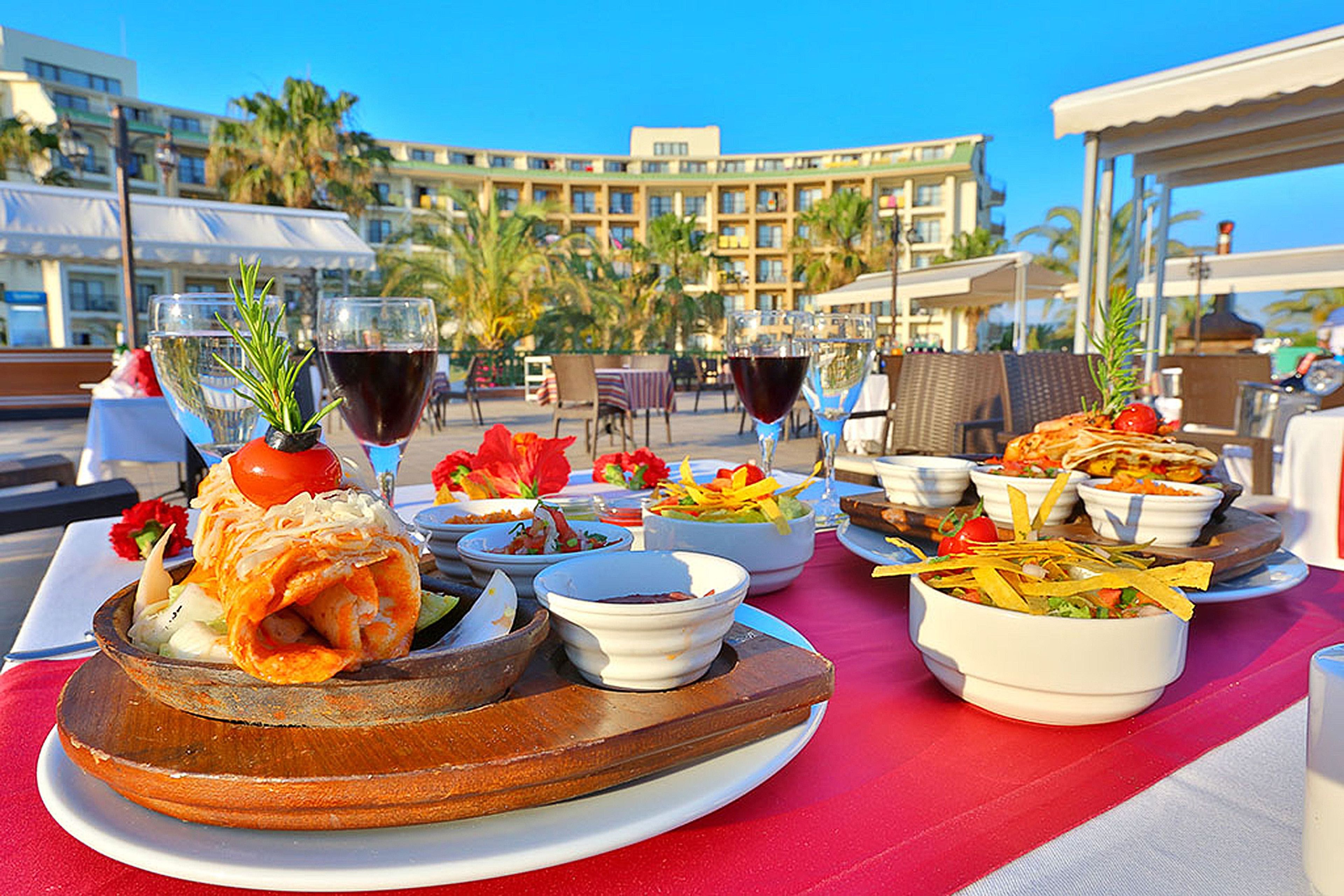 Mesa servida con diversos platos y bebidas, con vista a un hotel con palmeras y un cielo despejado.