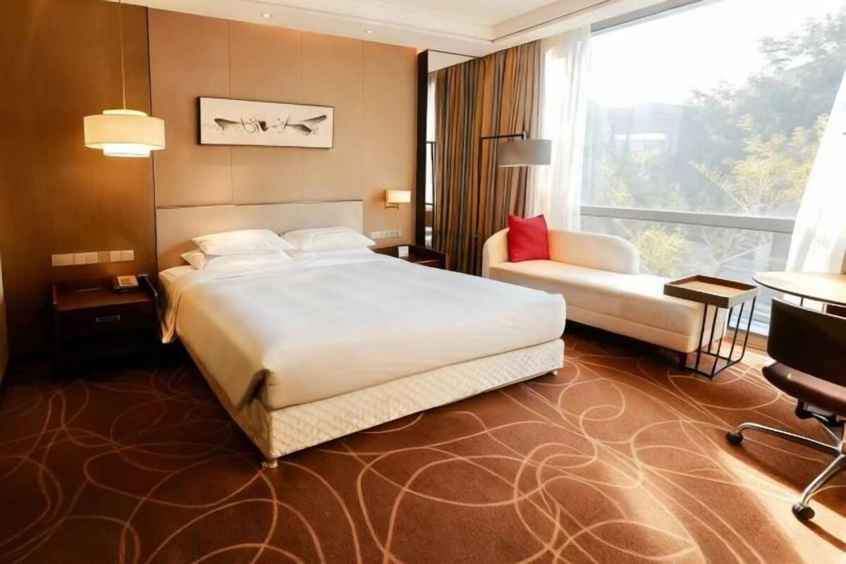 A hotel room with a large bed, a sofa, a desk, and a view of trees through the window.