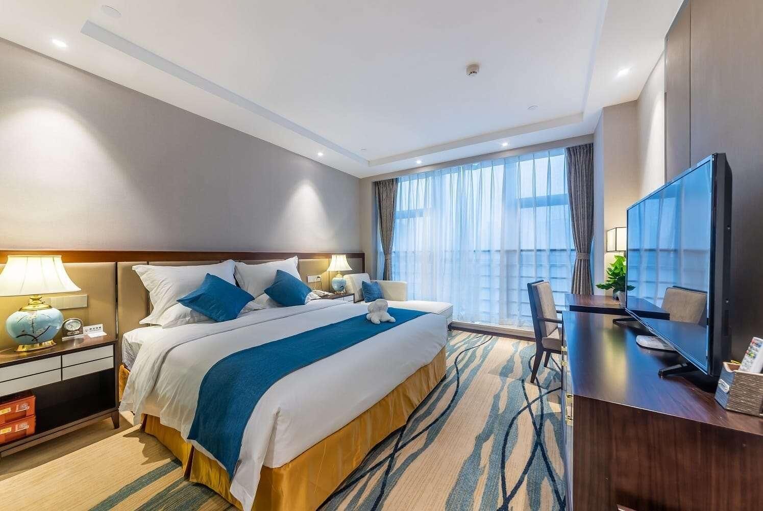 A hotel room with a large bed, a desk with a TV, and a view of the city through the window.