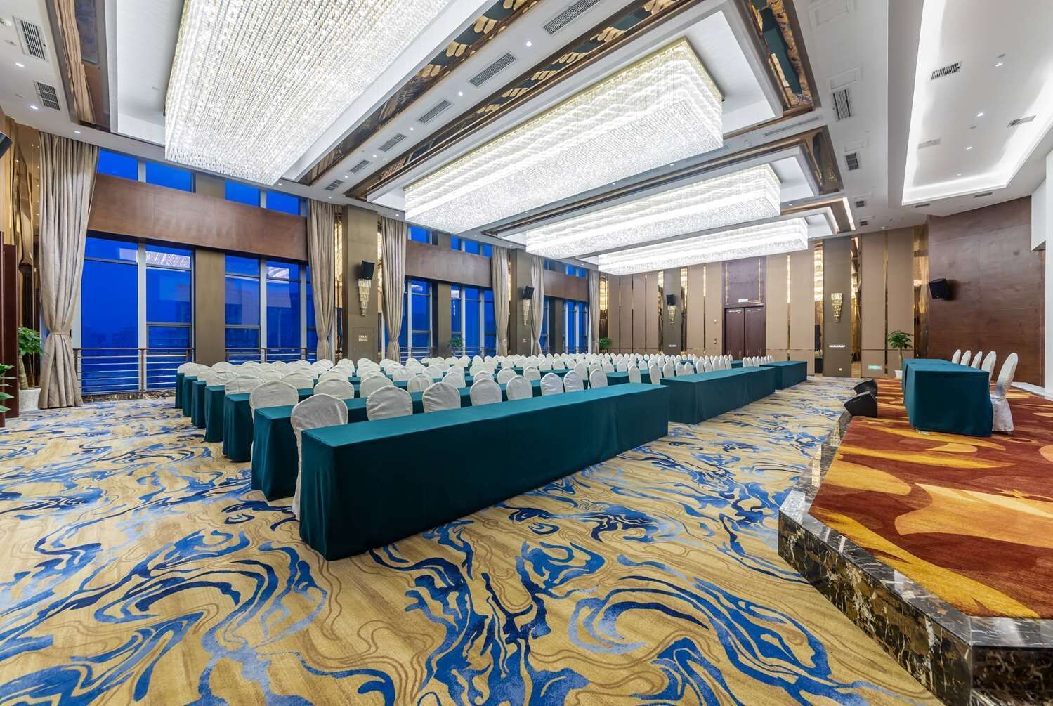 Une salle de conférence avec des rangées de tables et de chaises, de grandes fenêtres et un tapis à motifs.
