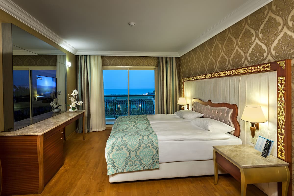 A hotel room with a large bed, a desk, a TV, and a balcony with a view of the sea.