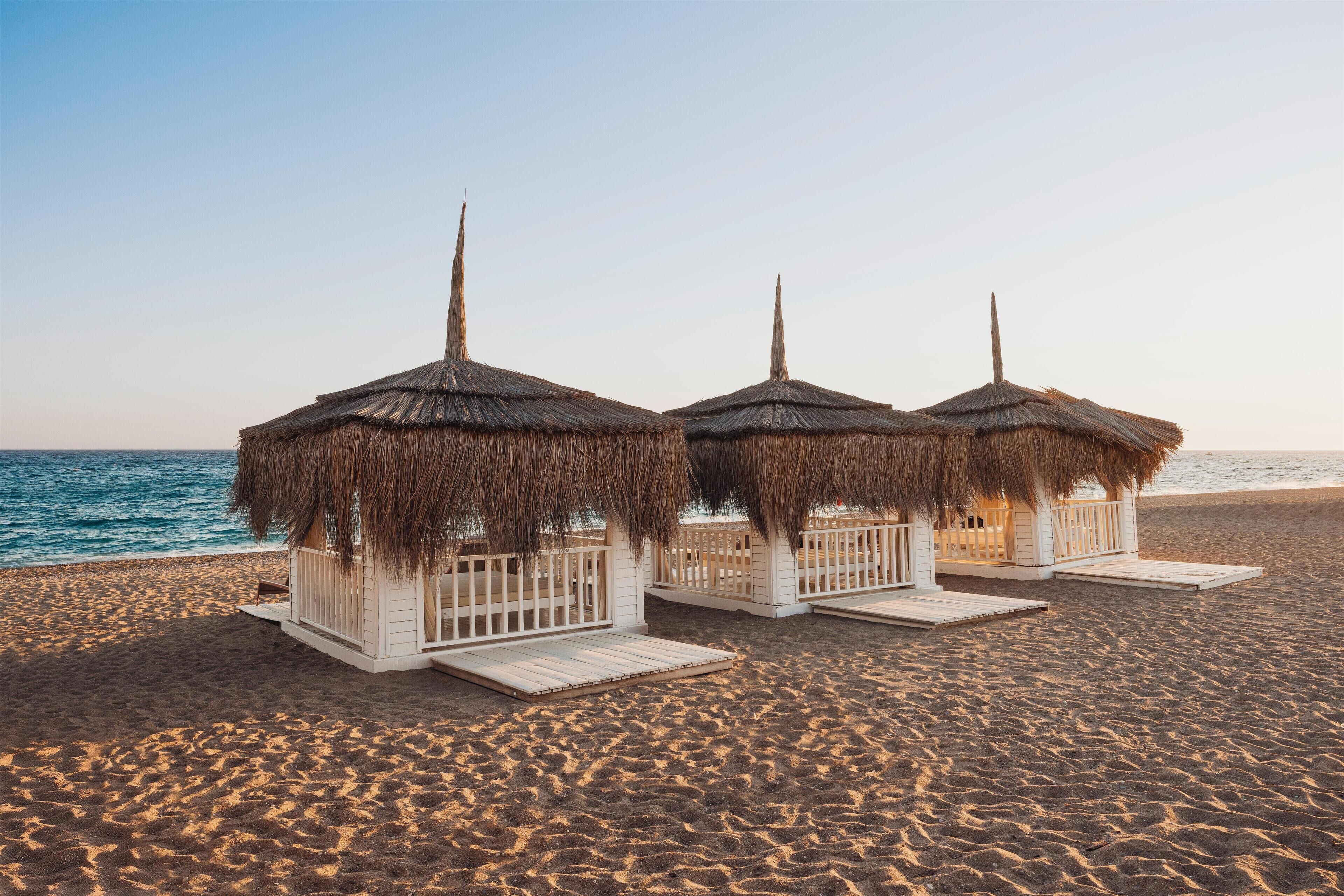 Tres cabañas de techo de paja frente a la playa, con barandillas de madera y escalones que dan al mar.
