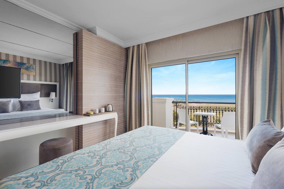 A hotel room with a bed, a desk, a chair, and a balcony with a view of the beach.