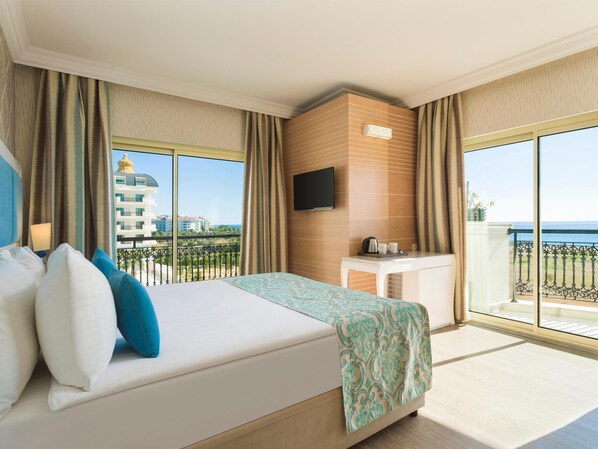 A hotel room with a large bed, a TV mounted on the wall, and a balcony with a view of the sea and buildings.
