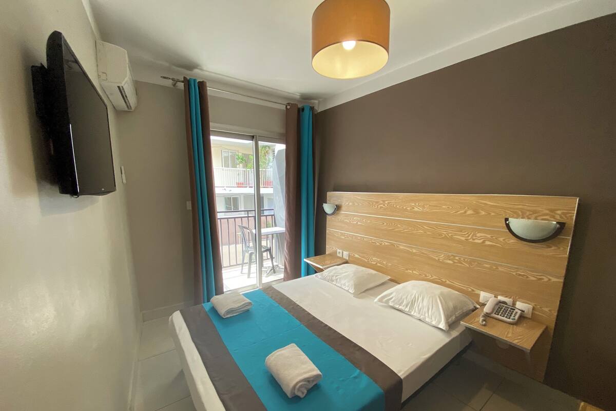 Habitación de hotel con cama, mesita de noche con lámpara, televisor montado en la pared y balcón con vistas al exterior.