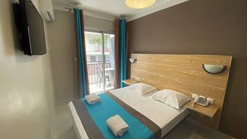 Una camera d'hotel con un letto, un comodino con una lampada, una TV a parete e un balcone con vista sull'esterno.