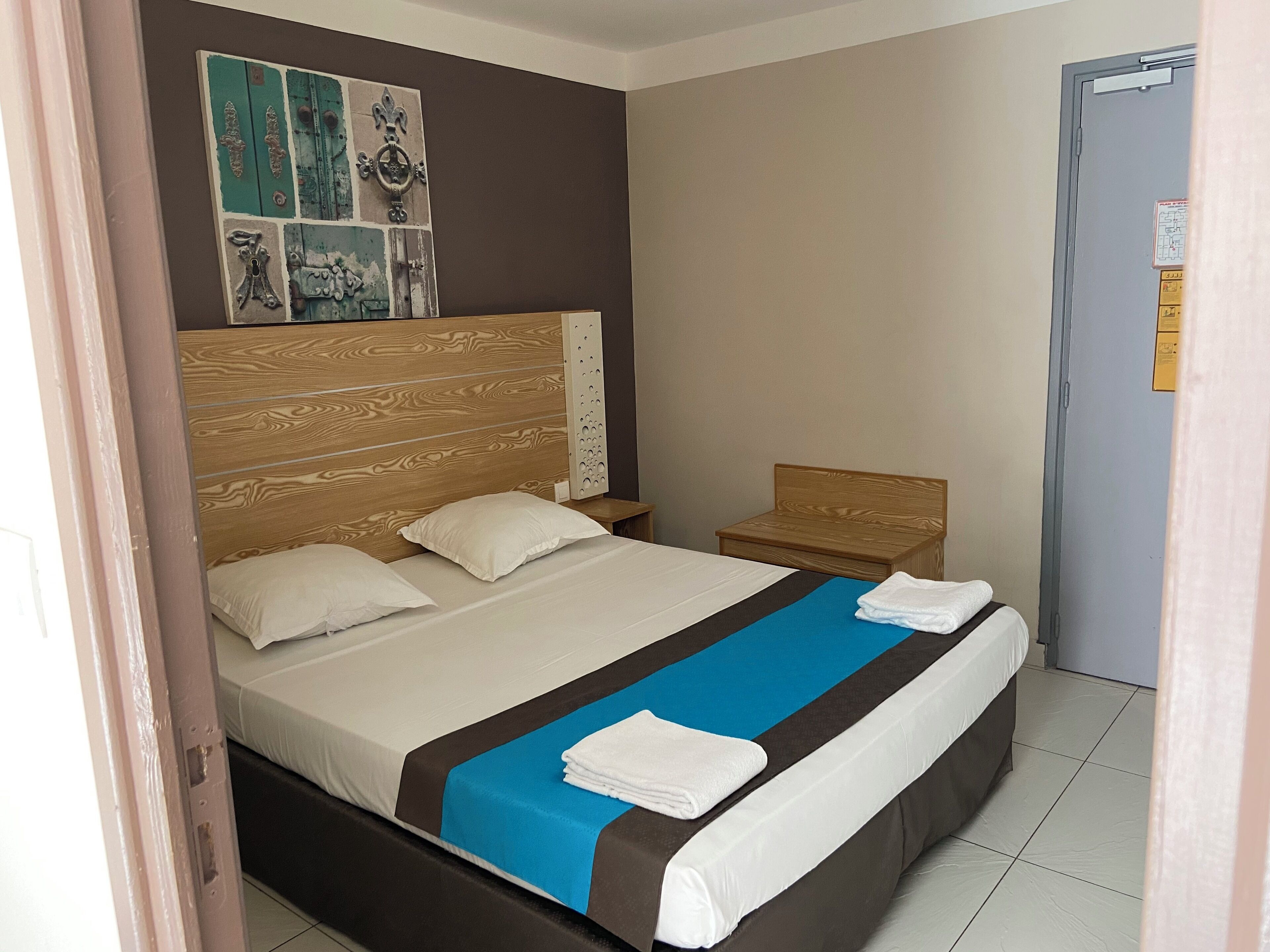 A hotel room with a wooden headboard, a bed with white and blue bedding, a small wooden nightstand, and a door with a map and instructions.