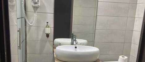 A modern bathroom with a glass shower enclosure, a white sink with a wooden vanity, a toilet, and a trash bin.