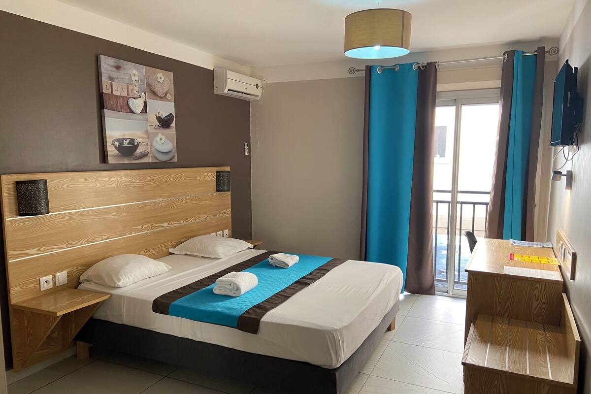 Habitación de hotel con cabecera de madera, cama con ropa blanca, toalla azul, escritorio con silla y ventana con cortinas.