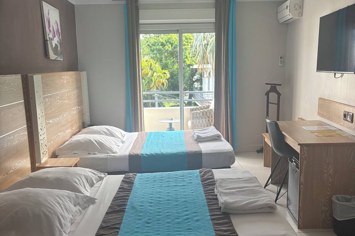 Habitación de hotel con dos camas, un escritorio, una silla y un balcón con vista a zonas verdes.