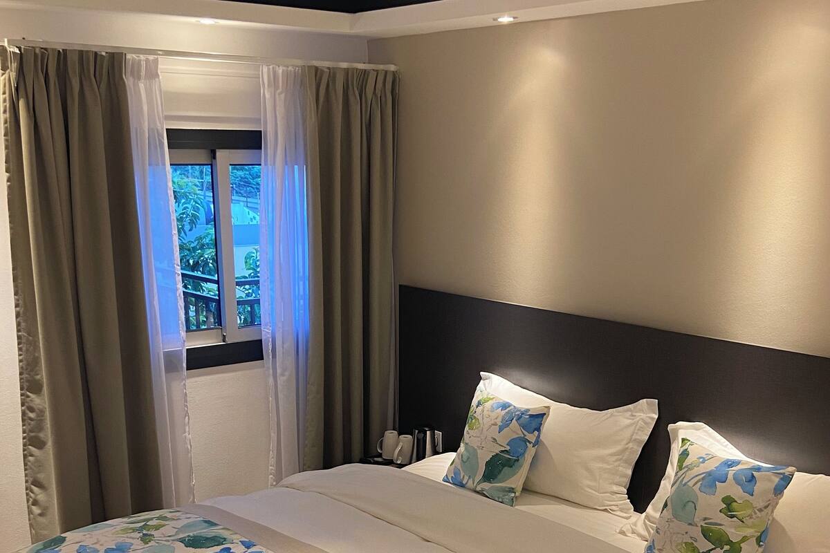 Un dormitorio moderno con una cama grande, cortinas color beige y vistas al exterior con vegetación.