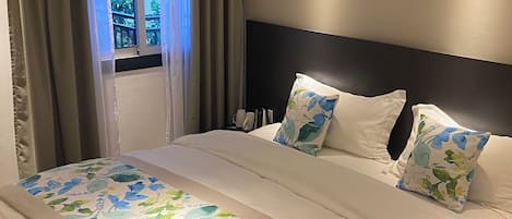 A modern bedroom with a large bed, beige curtains, and a view of greenery outside.