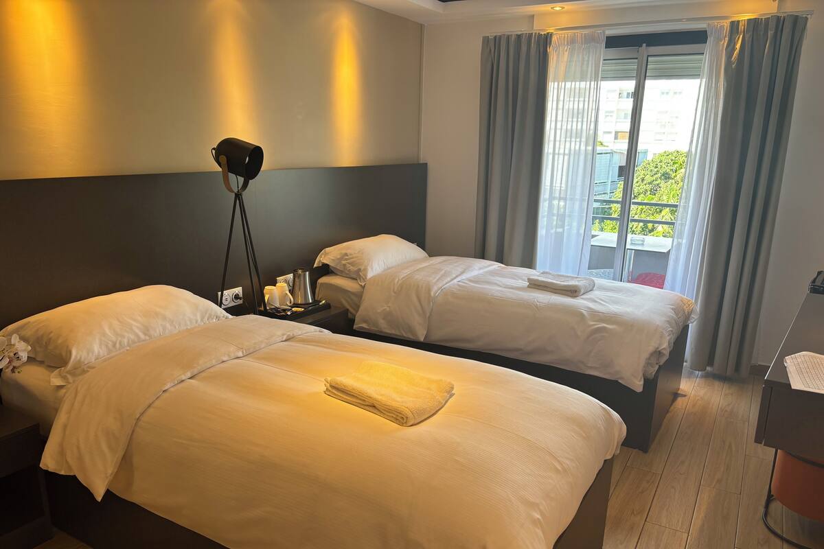 Habitación de hotel con dos camas, una mesita de noche con lámpara, una ventana con cortinas y vistas al exterior con vegetación.