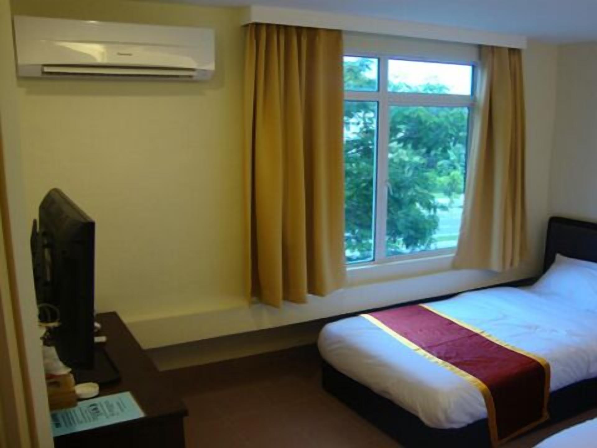 A hotel room with a bed, a desk with a computer, an air conditioner, and a window with curtains.