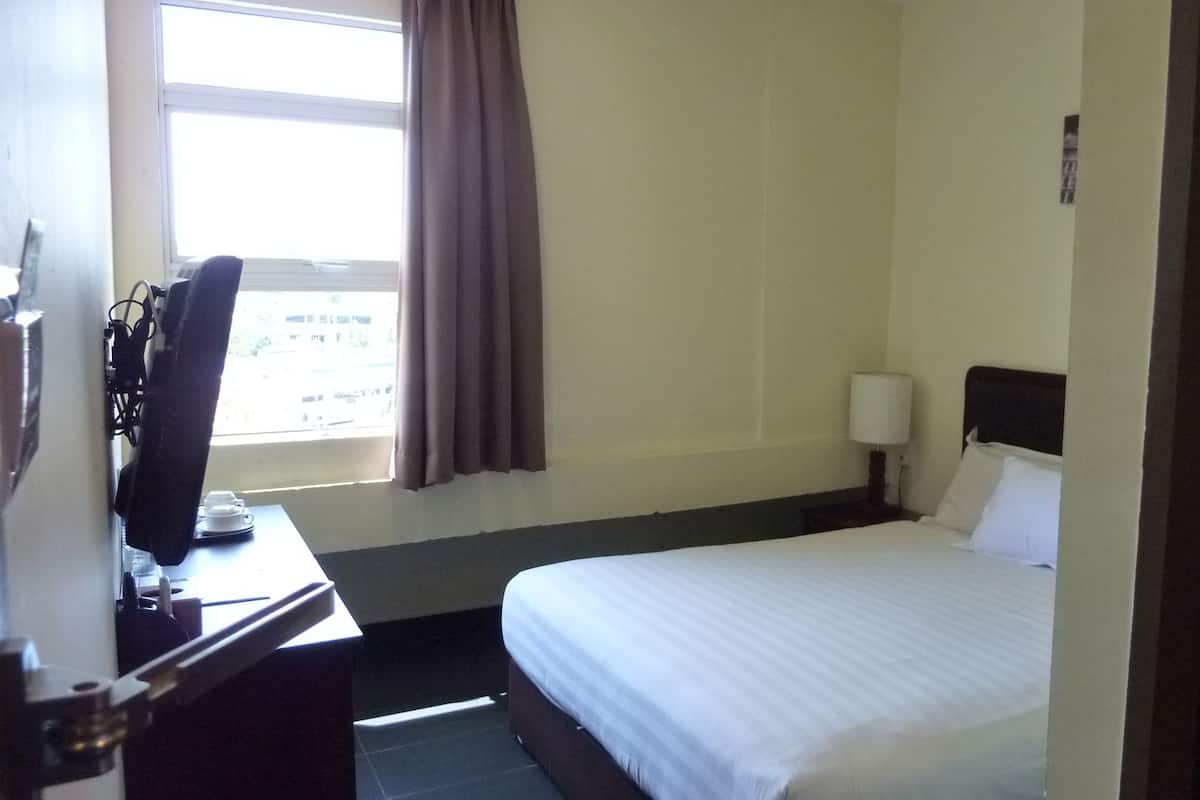 Una habitación de hotel con una cama, un escritorio con ordenador, una ventana con cortinas y una mesita de noche con lámpara.
