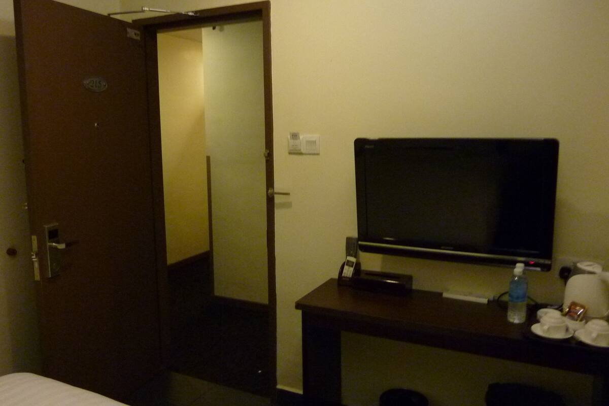 Habitación de hotel con una cama, un escritorio con televisor, una mesita con tazas y una botella, y una puerta que da a otra habitación.