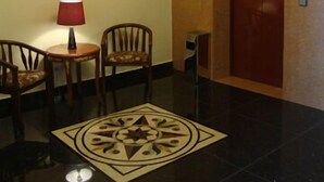 Um hall com porta de madeira, abajur vermelho, duas cadeiras, uma pequena mesa e um desenho estampado no chão.