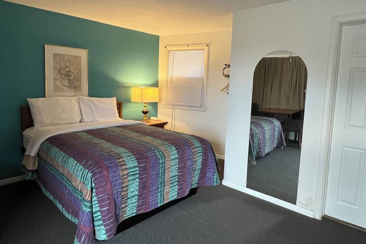 Una habitación de hotel con una cama, una lámpara de noche, un cuadro enmarcado en la pared y un espejo que refleja la habitación.