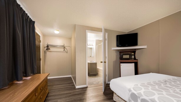 A hotel room with a wooden dresser, a bed with a patterned bedspread, a television on a shelf, and a bathroom visible through an open door. - Rodeway Inn La Crosse (La Crosse)