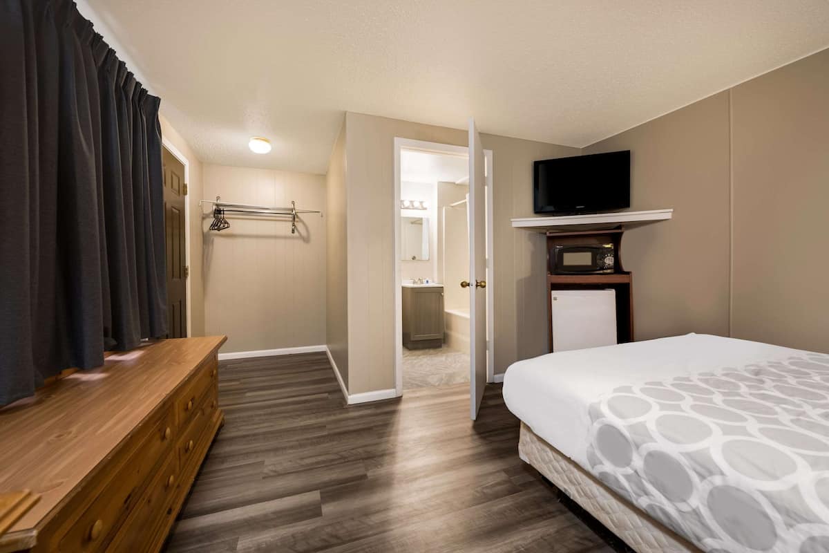A hotel room with a wooden dresser, a bed with a patterned bedspread, a television on a shelf, and a bathroom visible through an open door.