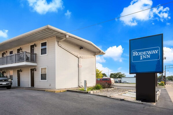 A two-story motel with a balcony on the second floor, a signboard for the motel, and a parking area with a few vehicles.