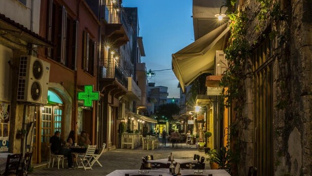 An outdoor dining area with tables and chairs, a green cross sign, and buildings with balconies. - Elia Daliani (Chania)
