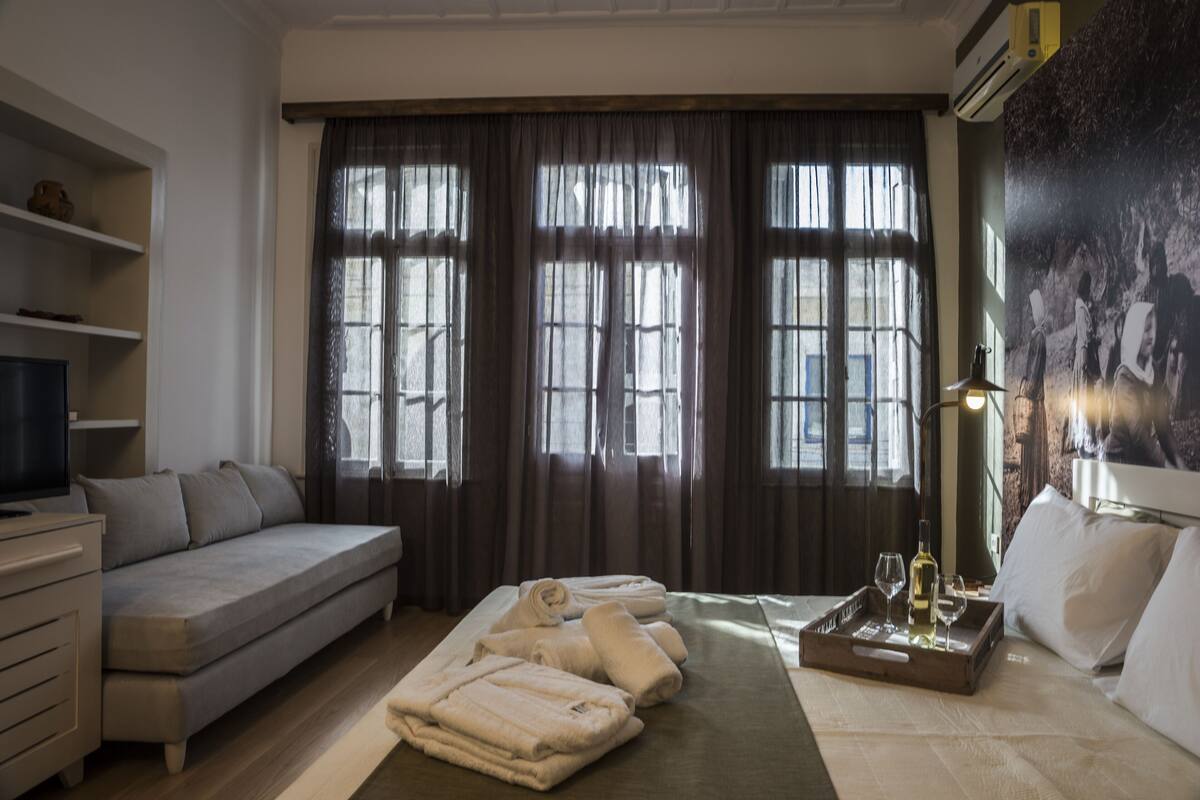 Et hotelværelse med sofa, seng, fjernsyn, bakke med håndklæder og vinglas samt store vinduer med gardiner.