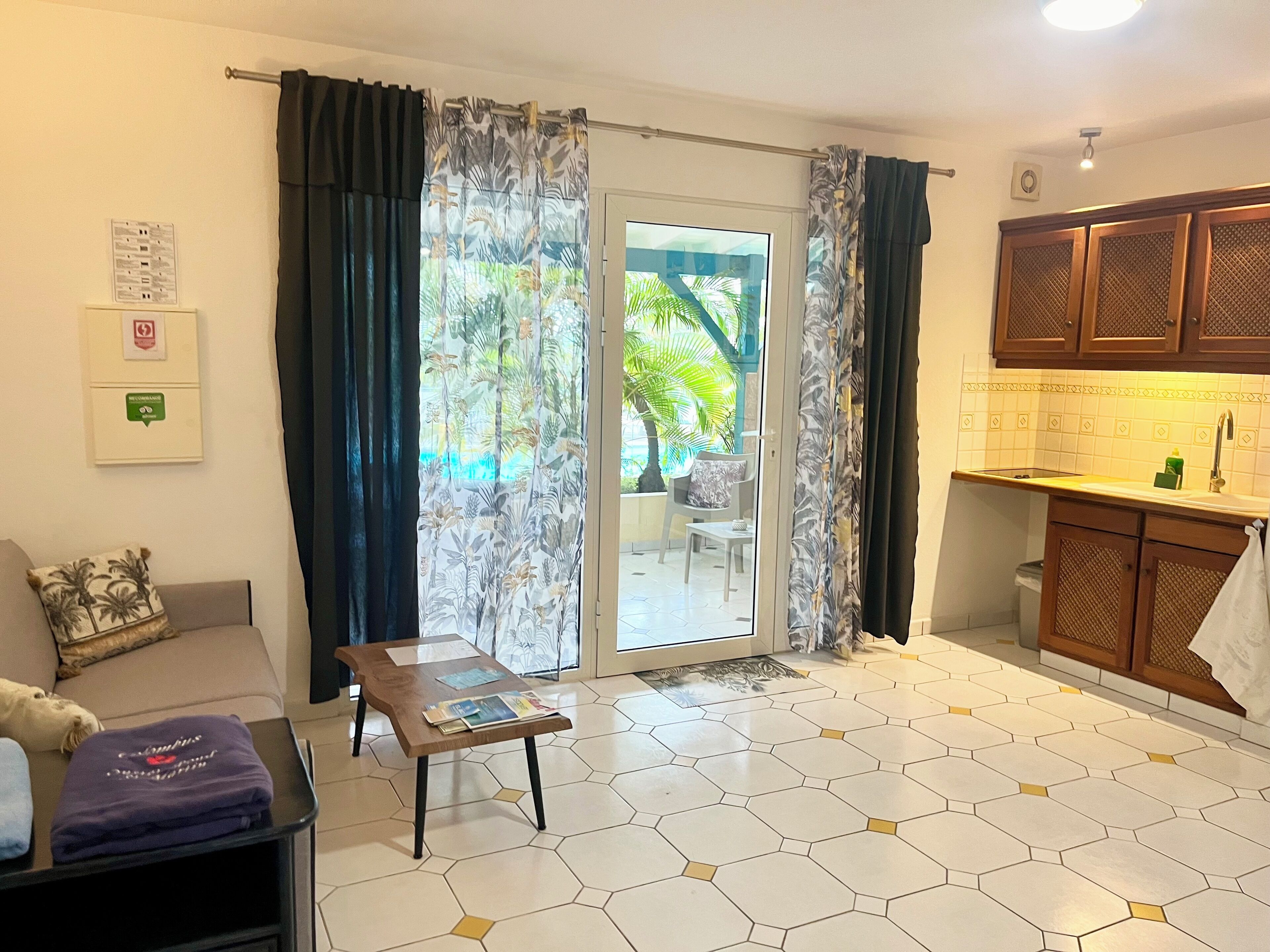 A living room with a sofa, a coffee table, and a view through a sliding glass door to an outdoor area with a pool and palm trees.