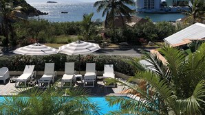A pool area with lounge chairs and umbrellas, a palm tree, and a view of the ocean and a building in the background. - Colombus Résidence (Oyster Pond)