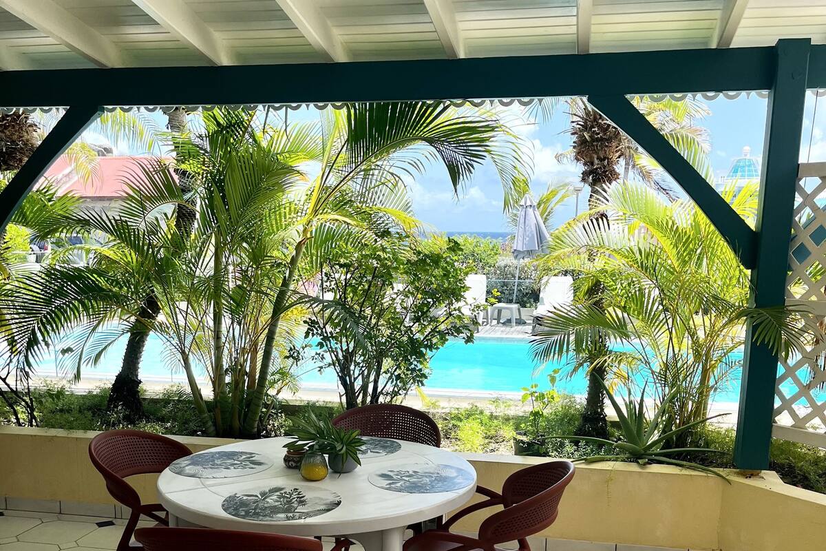 A covered outdoor seating area with a round table and chairs, surrounded by tropical plants and overlooking a pool.
